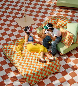 Two people sitting on colourful furniture under a large yellow umbrella on a checkered floor, surrounded by green and orange squishy cotton bedding.