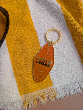 A retro-style keychain with a gold-tone metal ring and a tan-orange body, featuring the text 'WEIRDSTOCK' and 'Sweet Dreams' with moon and star graphics. The keychain is displayed on a yellow and white striped towel.