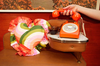 Colourful retro print heart cushion in green, pink, lilac, orange and red with ruffle edge next to a manicured hand holding a vintage orange telephone